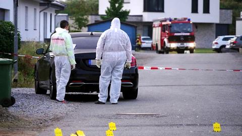 Zwei Personen in weißen Schutzanzügen stehen vor einem schwarzen Auto in einer mit Absperrband abgeteilten Straße. Im Bildvordergrund sind gelbe Marken auf dem Boden zu sehen.