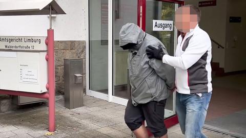 Ein Mann, dessen Kopf unkenntlich gemacht wurde, führt einen anderen Mann, dessen Kopf von der Kapuze der grauen Jacke verdeckt wird, aus einem Gebäude. Daneben das Schild "Amtsgericht Limburg".