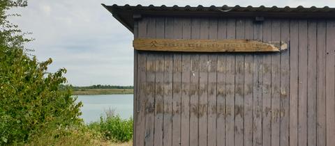 Vorne eine Hütte mit Schild "Pfaffensee und Teufelsee", im Hintergrund der See