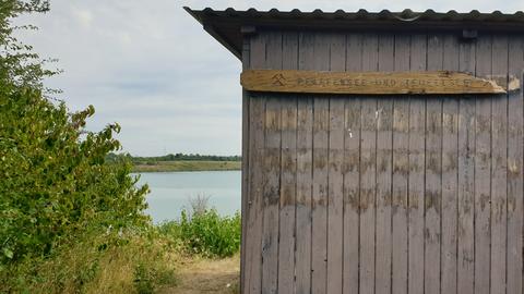 Vorne eine Hütte mit Schild "Pfaffensee und Teufelsee", im Hintergrund der See