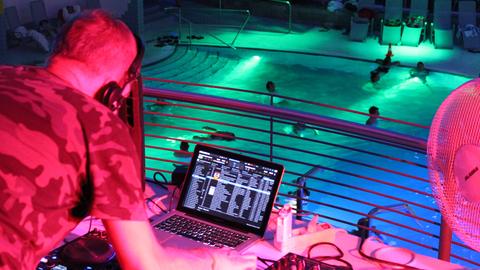 Im Bildvordergrund unscharf ein DJ am DJ-Pult während er Musik einspielt. Im Bildhintergrund scharf ein farbig beleuchtetes Schwimmbecken mit Menschen. 