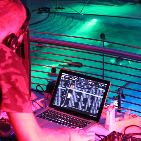 Im Bildvordergrund unscharf ein DJ am DJ-Pult während er Musik einspielt. Im Bildhintergrund scharf ein farbig beleuchtetes Schwimmbecken mit Menschen. 