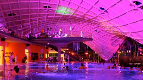 Der Innenraum des Gebäudes des Thermalbades farbig beleuchtet. Aus dem großen Becken ragt wie ein Pilz eine Bühne, auf welcher Musiker stehen.