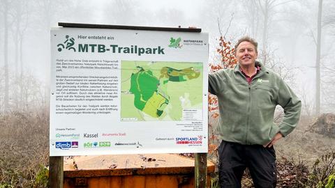 Ein Mann, Jürgen Depenbrock, steht neben einem großen Schild im Wald. Auf dem Schild steht "MTB Trailpark" mit Text und einer Karte