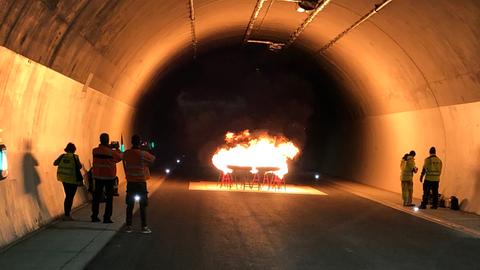 In einem Tunnel stehen Feuerschalen, aus denen helle Flammen lodern.