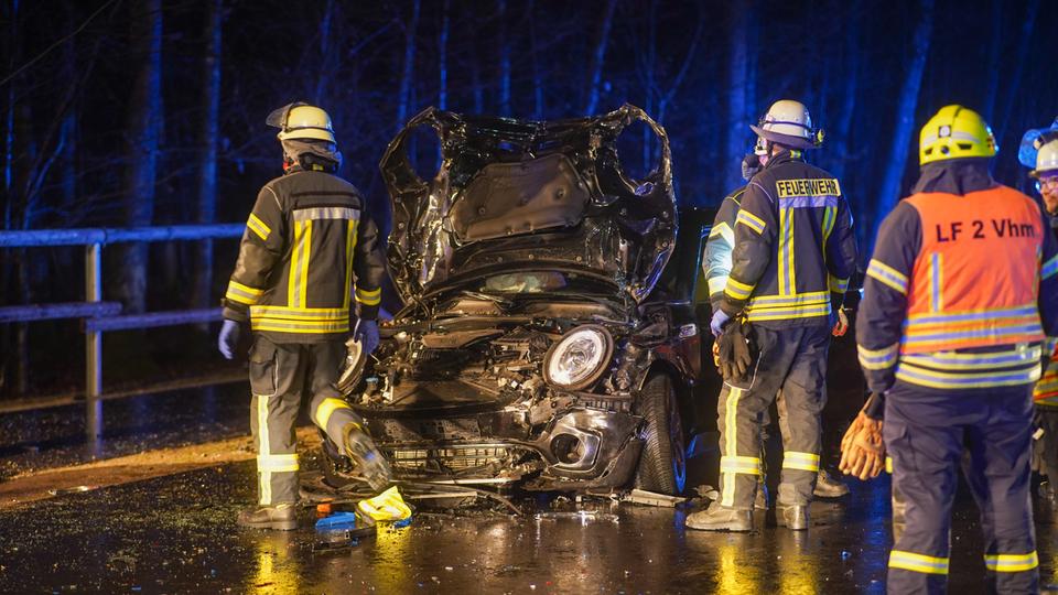 Das Unfallauto frontal fotografiert, die Motorhaube ist komplett zerstört. Drei Feuerwehrleute daneben. 