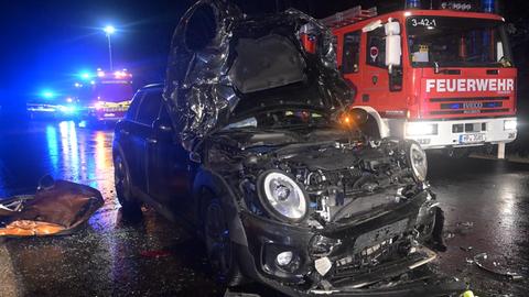 Das Unfallauto frontal fotografiert, die Motorhaube ist komplett zerstört. Drei Feuerwehrleute daneben. 