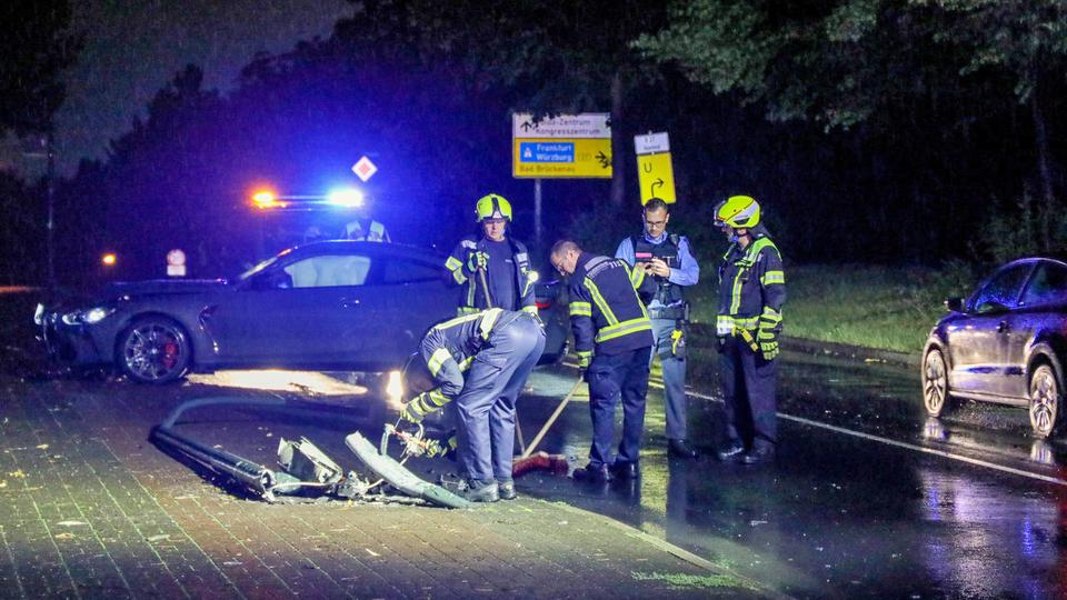 Unfall bei Nacht, Polizisten und Feuerwehrleute stehen an einem dunklen Auto, im Vordergrund ist eine umgeknickte Laterne zu sehen