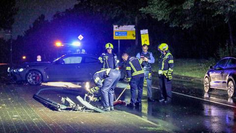Unfall bei Nacht, Polizisten und Feuerwehrleute stehen an einem dunklen Auto, im Vordergrund ist eine umgeknickte Laterne zu sehen