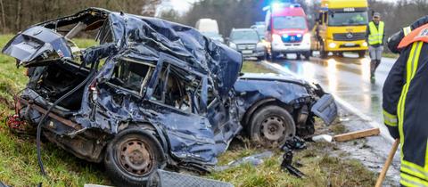 Völliges demolierter dunkler Pkw neben dem Fahrbahnrand, dahinter ADAC und Feuerwehr.