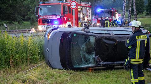 Ein Auto liegt neben einer Landstraße auf der Seite, daneben ein Feuerwehrmann und ein Feuerwehrauto.