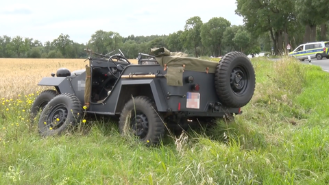 Ein Oldtimer-Allrad-Fahrzeug liegt neben der Straße, ein Reifen befindet sich neben dem Fahrzeug. Das Fahrzeug sieht aus wie ein alter Jeep - in dunkelgrau. 