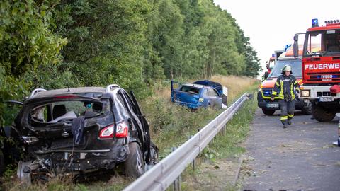 Zwei beschädigte Autos stehen im Gras neben einer Autobahn, rechts daneben Einsatzfahrzeuge und ein Feuerwehrmann.