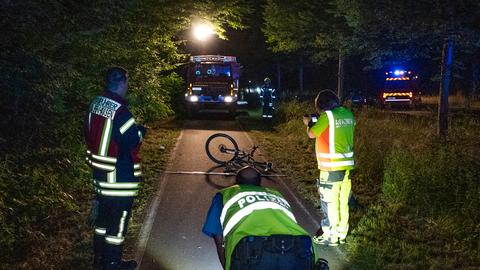 Drei Einsatzkräfte schauen auf die Unfallstelle auf einem Rad- und Fußgängerweg, auf dem ein Rad liegt. Dahinter steht ein Feuerwehrauto.