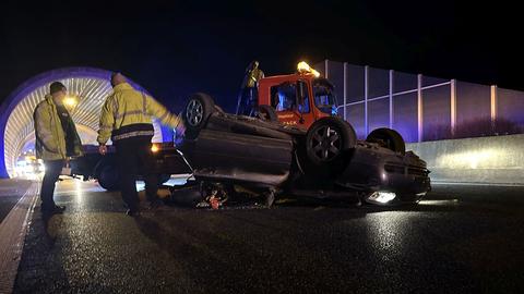 Das Auto liegt auf dem Dach, dahinter steht ein Feuerwehrauto. Zwei Rettungskräfte stehen daneben. 