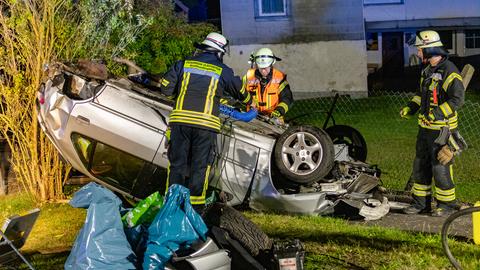 Rettungskräfte stehen an einem Auto, das auf einer Wiese auf dem Dach liegt.