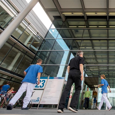Der Eingang der Uniklinik Frankfurt aus der Froschperspektive fotografiert. Mitarbeitende in Arbeitskleidung und Patientinnen und Patienten gehen ein und aus.