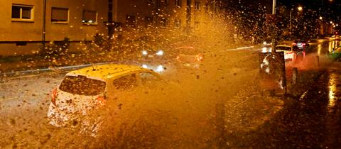 Autos fahren im Dunkeln in einer städtischen Straße durch eine große Pfütze, dabei spritzt Wasser in die Luft.