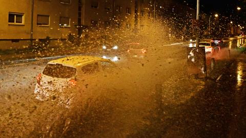 Autos fahren im Dunkeln in einer städtischen Straße durch eine große Pfütze, dabei spritzt Wasser in die Luft.