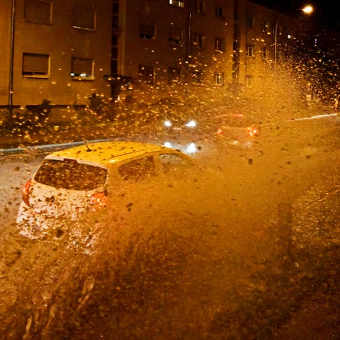 Autos fahren im Dunkeln in einer städtischen Straße durch eine große Pfütze, dabei spritzt Wasser in die Luft.