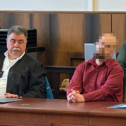 Ein Angeklagter in rotem Hemd sitzt zwischen seinen Verteidigern vor Gericht, sein Gesicht ist unkenntlich gemacht.