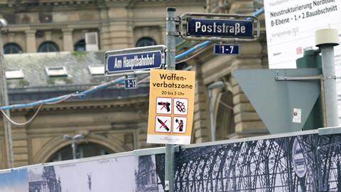 Schild mit der Aufschrift "Waffenverbotszone" und entsprechenden Symbolbildern hängt an einem Straßenschild mit den Namen "Am Hauptbahnhof" und "Poststraße".
