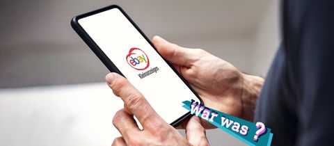 Hände halten ein Smartphone, auf dessen Screen das Logo von Ebay Kleinanzeigen zu sehen ist.