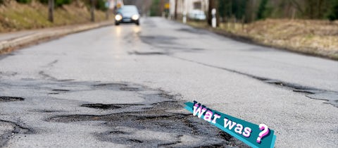 Foto einer Straße mit großflächigen Straßenschäden und Schlaglöchern in ländlicher Umgebung, im Hintergrund unscharf ein sich nahendes Auto.