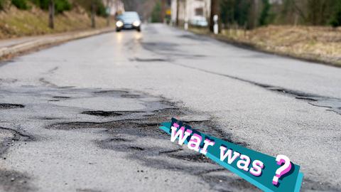 Foto einer Straße mit großflächigen Straßenschäden und Schlaglöchern in ländlicher Umgebung, im Hintergrund unscharf ein sich nahendes Auto.