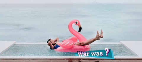 Foto eines Mannes, der in einem pinkfarbenen, aufgeblasenen Plastikschwan in einem Pool liegt und lächelnd den Regen auf sich niederprasseln lässt. Im Hintergrund das Meer. Auf dem Bild eine kleine, farbige Grafik mit dem Schriftzug "war was?".