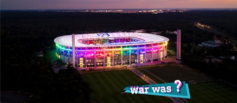 Foto vom Frankfurter WM-Stadion in der Abenddämmerung angestrahlt in Regenbogenfarben (Vogelperspektive). Auf dem Foto eine kleine farbige Grafik mit dem Schriftzug "War was?".
