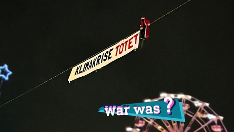 Eine Collage eines Weihnachtsmanns auf einem Drahtseil an dem ein Transparent hängt mit der Aufschrift "Klimakrise tötet" mit einer Weihnachtsmarkt Szene im Hintergrund.Liegt im Auf dem Bild eine kleine, farbige Grafik mit dem Schriftzug "war was?".