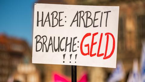 Während eines Warnstreiks wird ein Pappschild hochgehalten, auf dem "Habe: Arbeit - Brauche: Geld" steht.