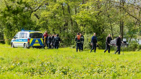 Eine Gruppe von Polizisten steht auf einer Wiese, im Hintergrund ein Fluss. Zwei Männer transportieren eine Trage mit einem Leichensack.