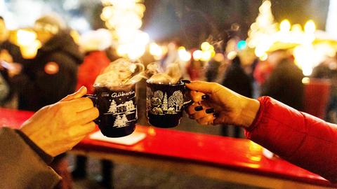 Im Vordergrund zwei Hände, die Tassen mit der Aufschrift "Glühwein" halten und anstoßen. Im Hintergrund unscharf das Treiben eines Weihnachtsmarktes.
