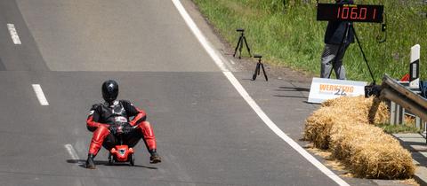 Der neue Weltrekordhalter sitzt auf einem gewöhnlichen roten Bobbycar, rechts auf einer Tafel werden 106 km/h angezeigt.