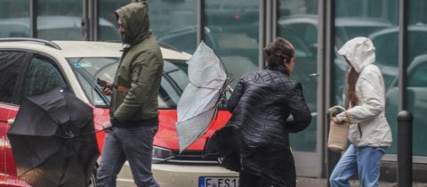 Menschen laufen in Frankfurt mit Regenschirmen über eine Straße - einige halten die Schirme quer, da es sehr windig ist. 