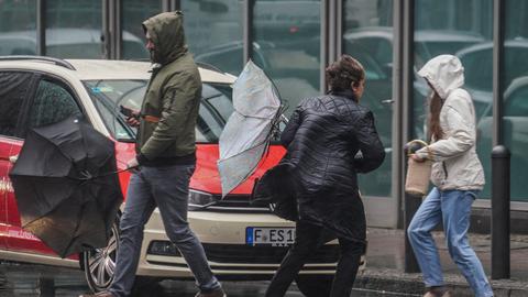 Menschen laufen in Frankfurt mit Regenschirmen über eine Straße - einige halten die Schirme quer, da es sehr windig ist. 