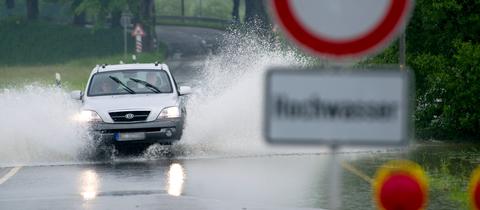 Ein Auto fährt durch Wasser, das in einer Straße steht. Im Bildvordergrund unscharf ein Schild "Achtung Hochwasser".