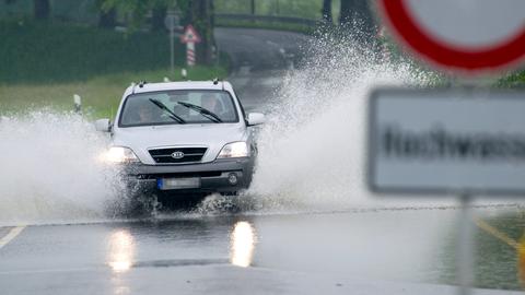 Ein Auto fährt durch Wasser, das in einer Straße steht. Im Bildvordergrund unscharf ein Schild "Achtung Hochwasser".