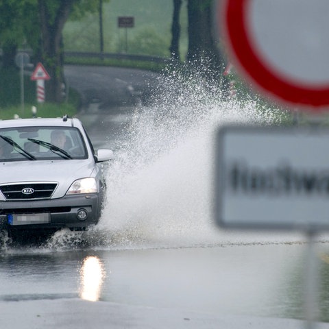 Ein Auto fährt durch Wasser, das in einer Straße steht. Im Bildvordergrund unscharf ein Schild "Achtung Hochwasser".