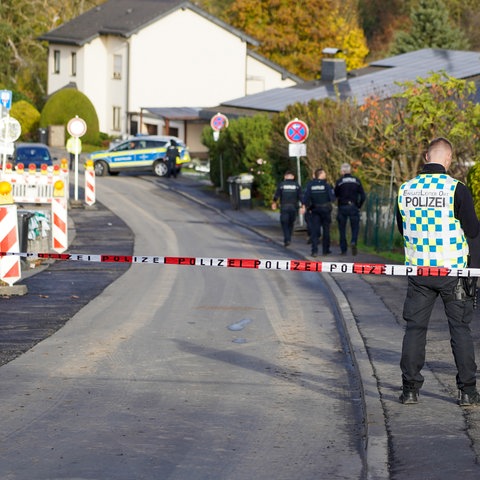 Blick in eine Straße eines Wohngebietes. Im Vordergrund ein Absperrband, hinter dem Absperrband Mitarbeitende der Polzei.