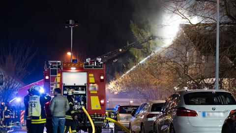 Totale. Eine nachtdunklestraße in Hofheim. Ausgeleuchtet von Scheinwerfern. Im Vordergrund die Rückseite eine Feuerwehrwagens und einige Feuerwehrleute. Im Hintergrund eine Feuerwehrleiter. Rechts davon ein rauchender Dachstuhl.