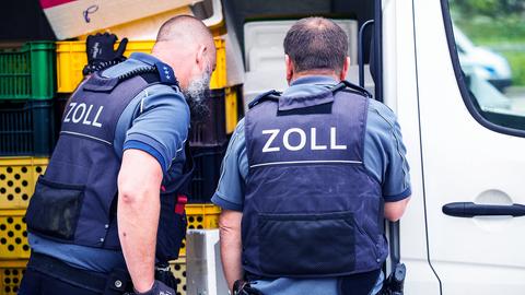Zwei Männer mit blauen Westen und der Aufschrift "Zoll" auf dem Rücken schauen in einen Lieferwagen voller Kisten.