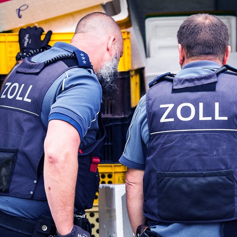 Zwei Männer mit blauen Westen und der Aufschrift "Zoll" auf dem Rücken schauen in einen Lieferwagen voller Kisten.