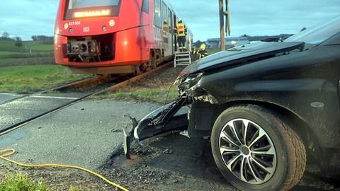 Das Bild zeigt ein schwarzes Auto mit beschädigter Front und einen roten Regionalzug. 