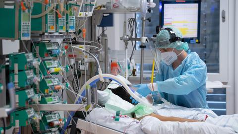 Viele Kabel und Schläuche an einem Bett, auf dem ein nicht zu erkennender Patient liegt, ein Pfleger in Schutzausrüstung daneben. 