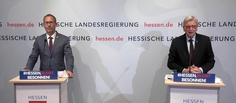 Gesundheitsminister Kai Klose und Ministerpräsident Volker Bouffier) stehen nebeneinander an Stehpulten. Im Hintergrund eine Wand mit den Schriftzügen und Logos der hessischen Landesregierung. 