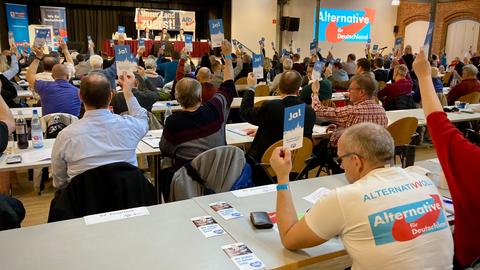 In einem Saal sitzen dutzende Menschen an Tischen und halten Abstimmungskarten hoch.