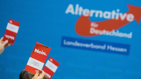 Parteilogo der AfD an einer blauen Wand. Davor in die Höhe gestreckte Hände mit "Nein"-Karten.